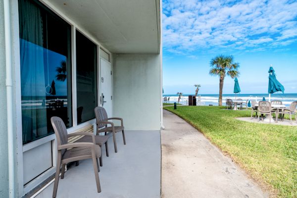 A beachfront patio with two chairs overlooks a path, lawn, palm tree, and distant ocean under a partly cloudy sky.