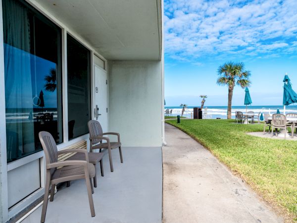 A beachfront patio with two chairs overlooks a path, lawn, palm tree, and distant ocean under a partly cloudy sky.