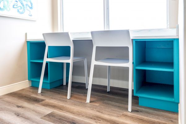 The image depicts a workspace with two white chairs at a white desk that has blue shelves underneath, situated in a brightly lit room.