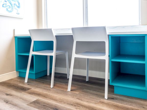 The image depicts a workspace with two white chairs at a white desk that has blue shelves underneath, situated in a brightly lit room.