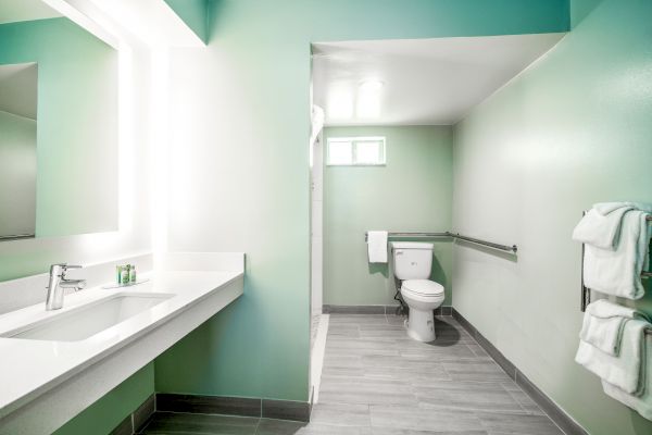 The image shows a modern bathroom with a long counter, sink, and mirror on the left, and a toilet with towels on racks to the right.