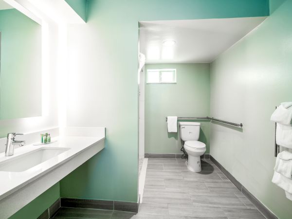 The image shows a modern bathroom with a long counter, sink, and mirror on the left, and a toilet with towels on racks to the right.