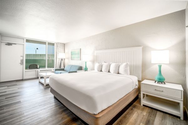 A modern, well-lit hotel room with a large bed, white linens, two nightstands, lamps, a sofa, a coffee table, and a view towards an outdoor area.