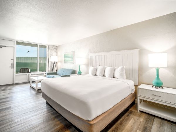 A modern, well-lit hotel room with a large bed, white linens, two nightstands, lamps, a sofa, a coffee table, and a view towards an outdoor area.