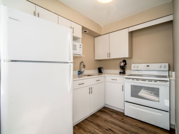 A small kitchen with white cabinets, a refrigerator, microwave, sink, countertop appliances, and a stove with an oven, all on wood flooring.