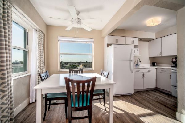 A bright kitchen and dining area with wooden flooring, white cabinets, a fridge, a white table with four chairs, window, and ceiling fan.