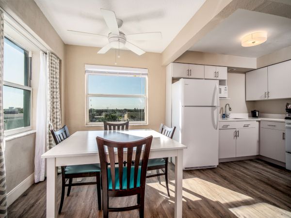 A bright kitchen and dining area with wooden flooring, white cabinets, a fridge, a white table with four chairs, window, and ceiling fan.
