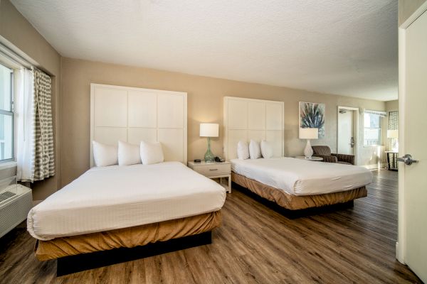 A hotel room with wood floors, two queen beds, a bedside table with a lamp, a chair, and a window with curtains. The room is well-lit and spacious.