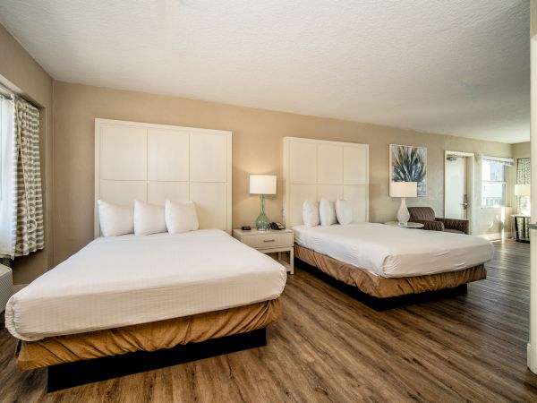 A hotel room with wood floors, two queen beds, a bedside table with a lamp, a chair, and a window with curtains. The room is well-lit and spacious.