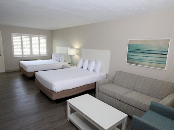 The image shows a hotel room with two beds, a sofa, an armchair, and a coffee table, featuring a serene beach painting on the wall.