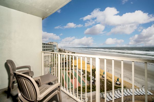 A balcony with two plastic chairs and a small table overlooks a sandy beach and ocean waves under a partly cloudy blue sky, with buildings in the distance.