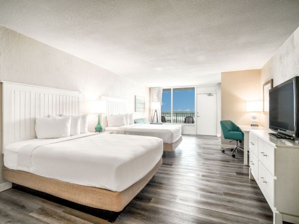 A modern hotel room with two double beds, a TV, desk, and chair. There's a balcony with a view of the ocean, creating a relaxing atmosphere.