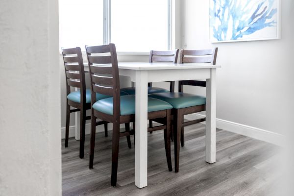 The image shows a dining area with a white table and four dark wooden chairs with teal cushions. A window and blue abstract painting are visible.