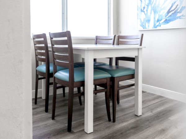 The image shows a dining area with a white table and four dark wooden chairs with teal cushions. A window and blue abstract painting are visible.