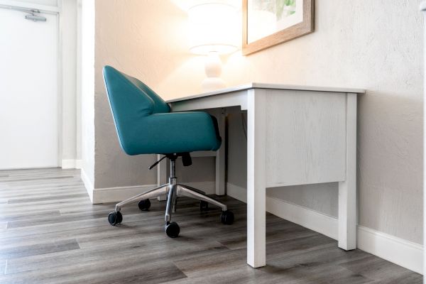 The image shows a modern, minimalist workspace with a white desk, a teal office chair on casters, a lamp, and a framed picture on the wall.