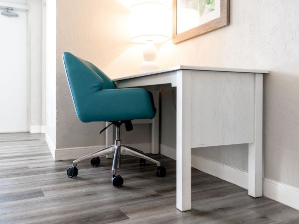 The image shows a modern, minimalist workspace with a white desk, a teal office chair on casters, a lamp, and a framed picture on the wall.