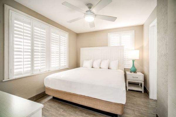 A cozy bedroom with a white bed, green lamp on nightstand, ceiling fan, and shutters on windows and door. The room has a bright, minimalist style.