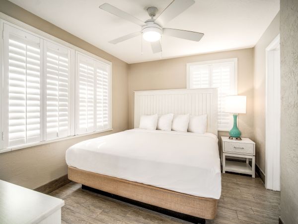 A cozy bedroom with a white bed, green lamp on nightstand, ceiling fan, and shutters on windows and door. The room has a bright, minimalist style.