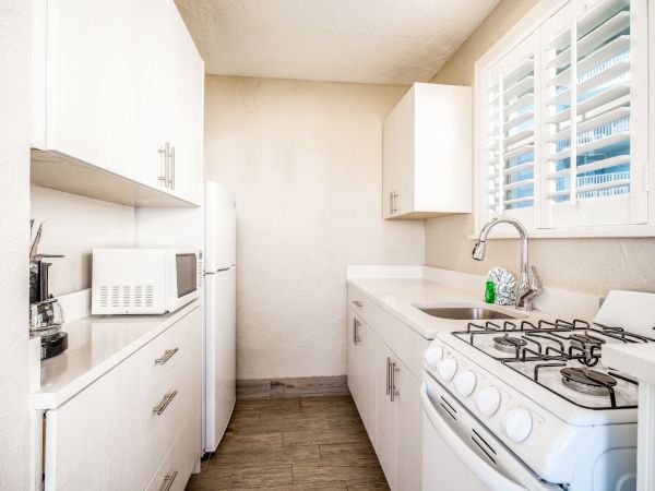 A small, bright kitchen with white cabinets, a gas stove, microwave, refrigerator, and a sink under a window with blinds.