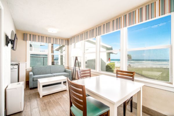 A bright, coastal-themed room with large windows showing a beach view, a dining table and chairs, a sofa, a lamp, and a mounted TV on the wall.