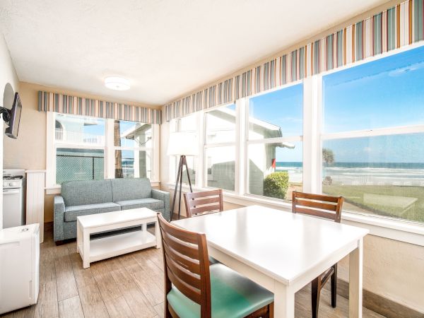 A bright, coastal-themed room with large windows showing a beach view, a dining table and chairs, a sofa, a lamp, and a mounted TV on the wall.