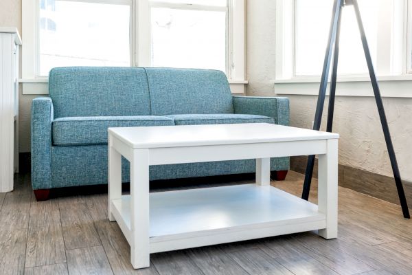 A modern living room features a blue sofa, a white coffee table, and a black tripod stand, with large windows letting in ample light.