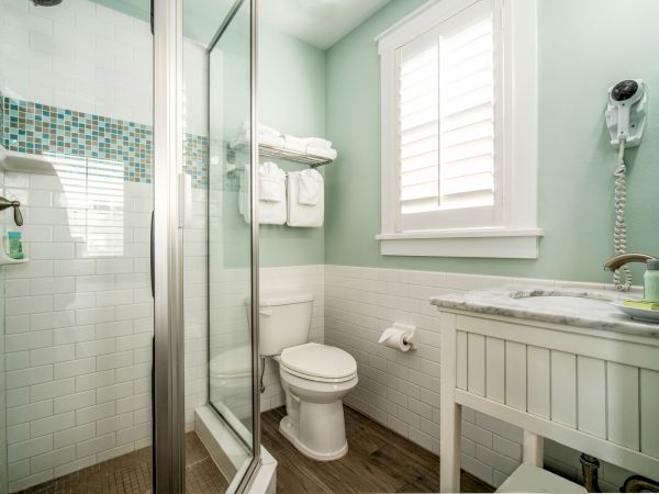 This image shows a modern bathroom with a glass shower, toilet, sink with countertop, wall-mounted hairdryer, and towels on a shelf, all in a pastel green.
