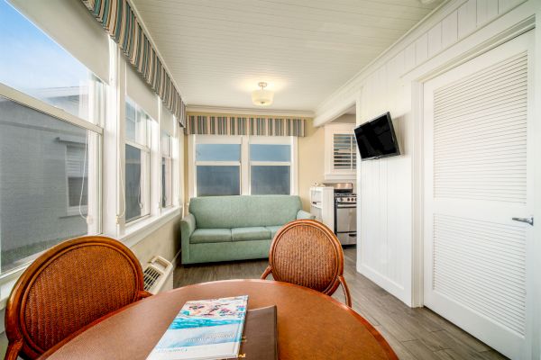 A bright room with large windows, a table and chairs, a sofa, a wall-mounted TV, and a small kitchen area with an oven and cabinets.