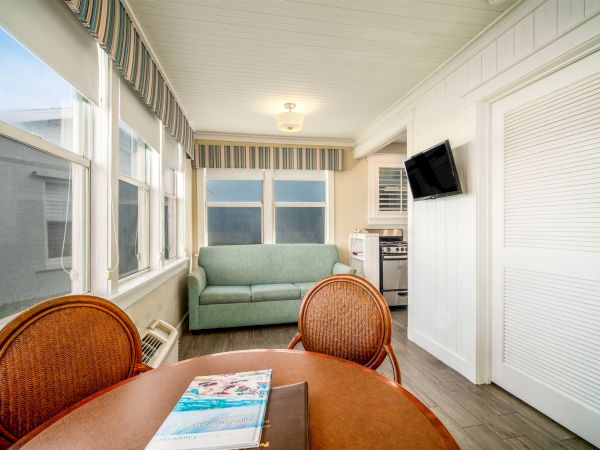 A bright room with large windows, a table and chairs, a sofa, a wall-mounted TV, and a small kitchen area with an oven and cabinets.