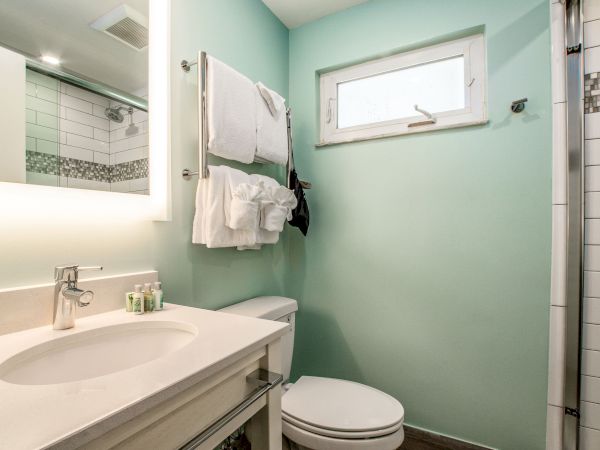 This image shows a modern bathroom with a light green wall, a sink with a mirror, a towel shelf, a toilet, and a shower area on the right.
