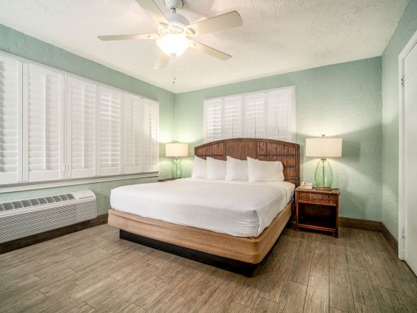 A modern bedroom with a large bed, two glass bedside lamps, a ceiling fan, green walls, wooden flooring, and white shutters on the windows ends the sentence.