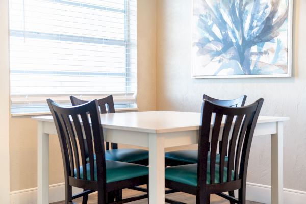 A small dining area with a white table, four black wooden chairs, a window with blinds, and a painting on the wall ending the sentence.