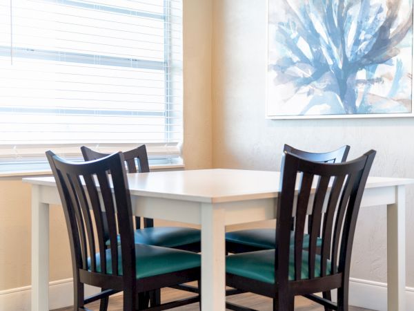 A small dining area with a white table, four black wooden chairs, a window with blinds, and a painting on the wall ending the sentence.