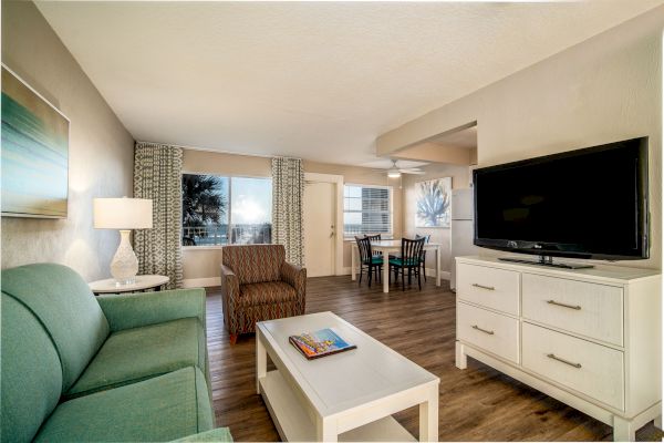 A cozy living room with a green couch, armchair, coffee table, TV on a white dresser, and a dining area with a table and chairs in the background.