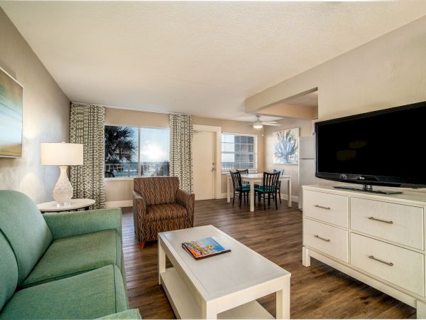 A cozy living room with a green couch, armchair, coffee table, TV on a white dresser, and a dining area with a table and chairs in the background.