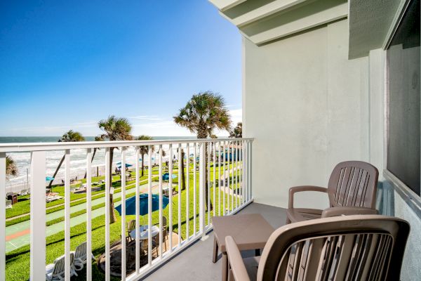 A balcony overlooking a lawn, palm trees, a pool, and the ocean, with two plastic chairs and a small table on the balcony.