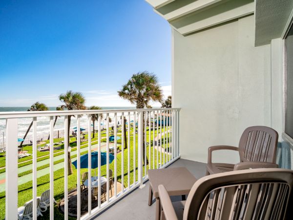 A balcony overlooking a lawn, palm trees, a pool, and the ocean, with two plastic chairs and a small table on the balcony.
