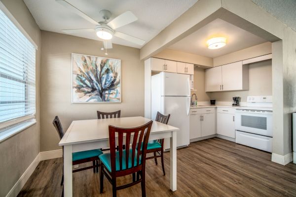 A modern kitchen with white cabinets, a dining table with four chairs, a refrigerator, an oven, a ceiling fan, and a colorful painting on the wall.