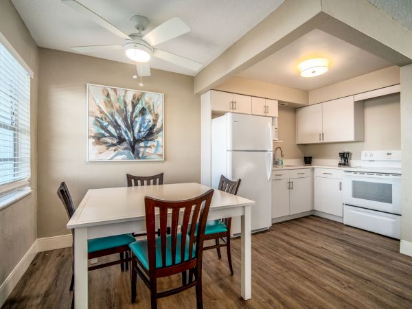 A modern kitchen with white cabinets, a dining table with four chairs, a refrigerator, an oven, a ceiling fan, and a colorful painting on the wall.