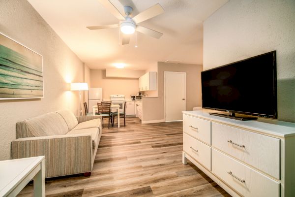 A modern living area with a sofa, TV, ceiling fan, and a dining table, leading to a kitchen in the background, all set on wooden flooring.