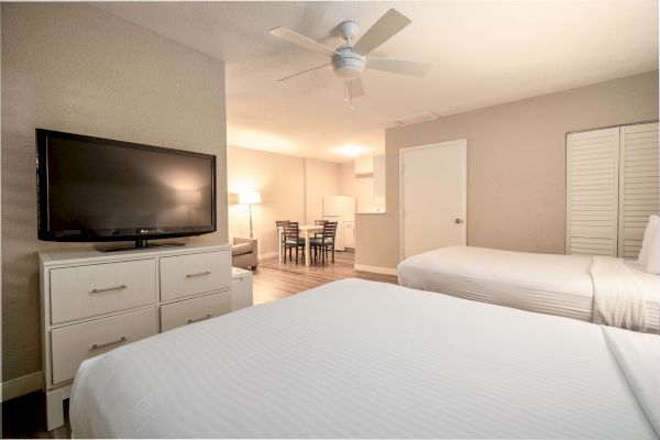 A hotel room with two beds, a flat-screen TV, a ceiling fan, a small dining area, and a kitchenette in the background.