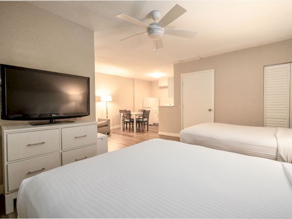 A hotel room with two beds, a flat-screen TV, a ceiling fan, a small dining area, and a kitchenette in the background.