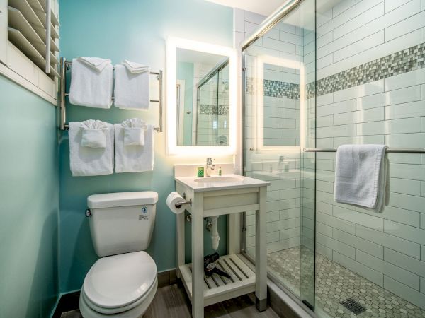 A modern bathroom with a toilet, sink, mirror, and a glass shower stall. Towels are neatly hung above the toilet and on the shower door.