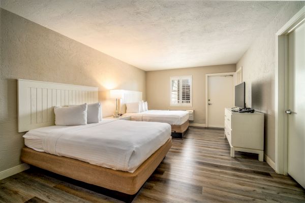 A clean, well-lit hotel room with two neatly made beds, a wooden floor, flat-screen TV on a dresser, and a window with blinds.