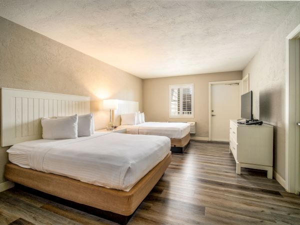 A clean, well-lit hotel room with two neatly made beds, a wooden floor, flat-screen TV on a dresser, and a window with blinds.
