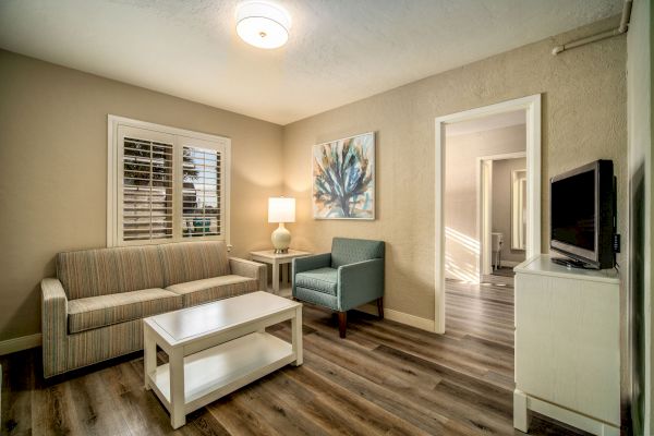 A cozy living room with a sofa, armchair, coffee table, lamp, wall art, TV, and wooden floor. An open door leads to another room.