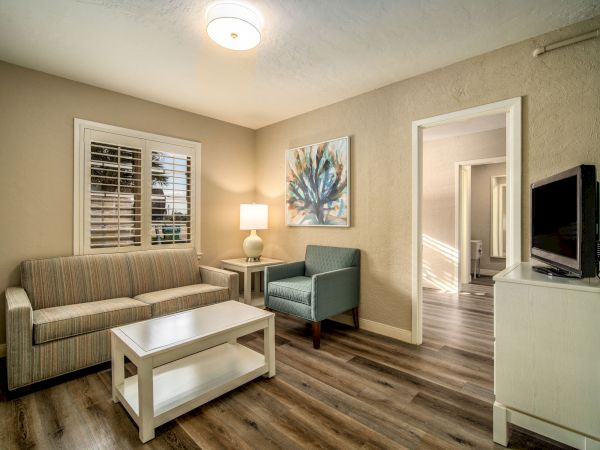 A cozy living room with a sofa, armchair, coffee table, lamp, wall art, TV, and wooden floor. An open door leads to another room.