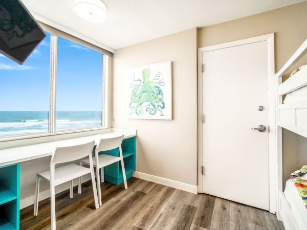 A room with a beach view, featuring a desk with two chairs, a bunk bed, and an octopus painting on the wall, all in a coastal-themed decor.