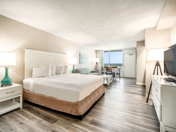 The image shows a modern hotel room with a king-sized bed, bedside tables with lamps, a TV, a sofa, a dining area, and a balcony view.