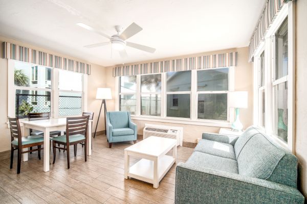 A bright living room with large windows, a ceiling fan, a dining table with chairs, a lounge chair, a sofa, a coffee table, and floor lamps.